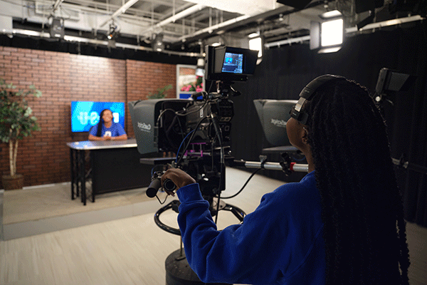 Inside a media newsroom, a Black student with long black dreadlocks stands behind a black camera and teleprompter, wearing a blue sweatshirt. On the set is a Black student wearing a blue T-shirt, blurred in the photo. A desk and television are visible on the set. A brick wall is in the background. Ceiling lights are also visible in the newsroom. 