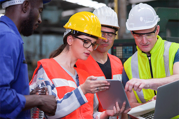 Diverse group of workers wearing hard hats and safety vests look at data on a tablet in a factory setting. 