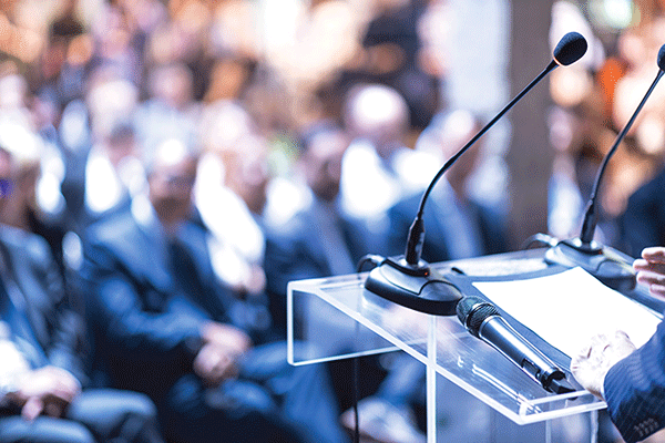 Detailed image of a podium with microphones set up in the left of the frame. A speaker’s hands are visible at the far left. An out-of-focus audience of business professionals is visible in the background.