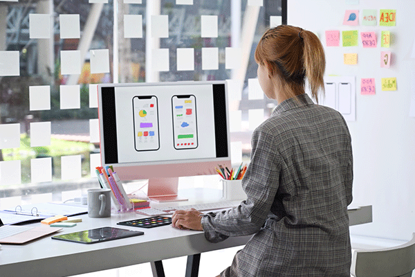 Router_design – A female with her back to the camera sitting in front of a large computer monitor looking at a graphic design with Post-It notes posted on a large window in front of the screen.