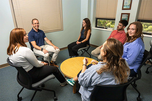 A group of six smiling individuals, a mix of men and women, sits chatting around a small yellow table in a cozy treatment space. They are dressed casually in a variety of styles and colors. There are two windows to the left where greenery can be seen beneath the partially drawn blinds. A larger window to the right has the blinds fully drawn.